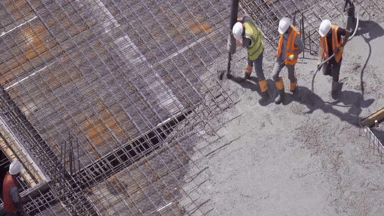 Drei Bauarbeiter in Warnwesten und Schutzhelmen verteilen frischen Beton auf einer großen Stahlbewehrung. Sie halten einen Betonschlauch und arbeiten gemeinsam an einer Baustelle, aus der Vogelperspektive aufgenommen.

                        